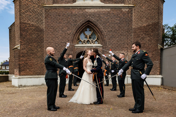 bruidspaar geeft kus voor de kerk in gassel en wordt omringd door reservisten met sabels