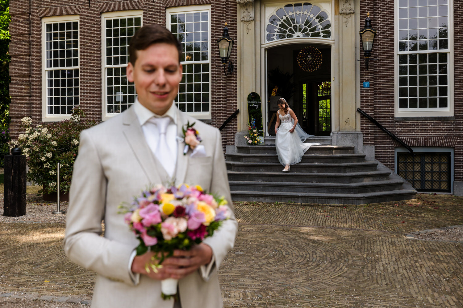 bruidegom op de voorgrond wacht op zijn bruid die vanuit het huis van buitenplaats sparrendaal naar hem toe loopt