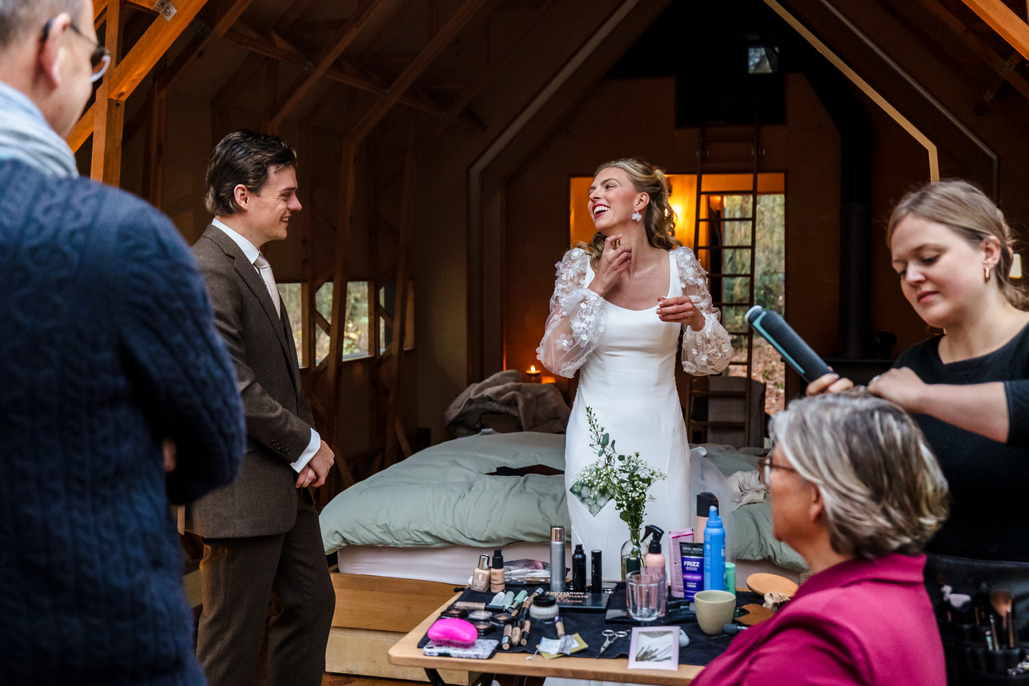 bruid doet luchtje op terwijl bruidegom toekijkt en moeder in de make-up zit